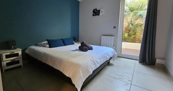 appart hôtel Pasino n°8 Chambre 1 tons dominante de bleus, au mur une décoration "sea". La chambre est composée d'un grand lit et ses 2 chevets blancs à tiroirs gris et leur lampe. Sur le lit, 2 coussins de décoration bleuet, 2 serviettes grises posées dessus. A l'arrière une porte fenêtre et son rideau occultant bleu un patio et ses palmiers