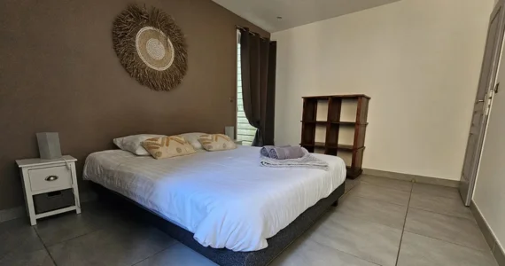 Appart hôtel Pasino n°7 chambre avec grand lit double 160x200 donnant sur un petit patio. Décoration dans les tons marrons avec deux coussins de décoration sur le lit, un plaid et 2 serviettes de bain. Chambre entièrement carrelée au sol (couleur grise). De chaque côté du lit, une table de chevet et sa lampe. La porte donnant sur le patio possède un rideau occultant. à droite un meuble de décoration et rangement en teck Au mur une décoration exotique.