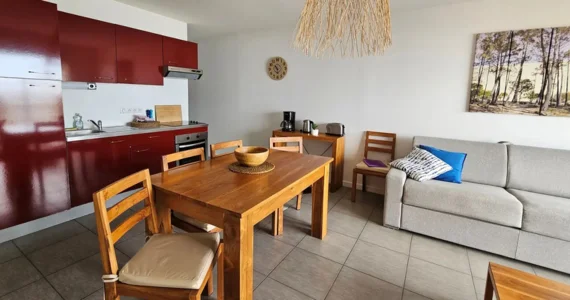 Appart hôtel Pasino n°5 pièce principal à gauche kitchenette rouge, au milieu une table en teck et ses chaises, au fond une console en teck sur laquelle sont posés une cafetière, un grille pain et une bouilloire, à droite un canapé et ses coussins. Au mur un tableau et une pendule