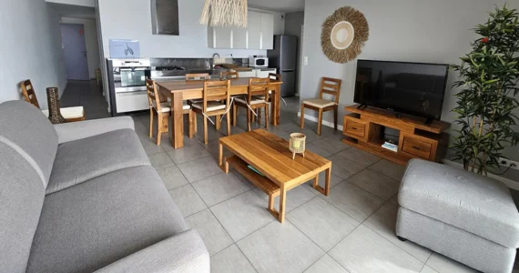 Appart hôtel Pasino n°6 pièce principale , un canapé gris, des meubles en teck, un écran TV, au milieu une table en teck et ses 8 chaises, au fond la cuisine équipée de meubles beiges et inox. Présence d'éléments de décoration aux murs et plafond. A droite un pouf et un palmier