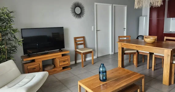 Appart hôtel Pasino n°5 pièce à vivre Au fond, la kitchenette équipée rouge, au milieu une table en teck et ses chaises. Sur la gauche à l'avant un fauteuil tournant blanc, une table basse en teck posé dessus un élément de décoration en verre bleu. Le long du mur au fond,, un palmier, un meuble TV et sa TV grand écran. Sur le mur une décoration exotique
