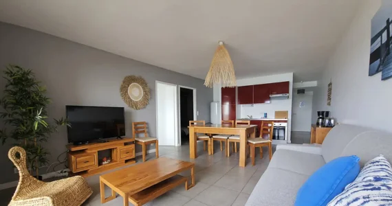Appart hôtel Pasino n°2 pièce à vivre de la résidence Pasino Au fond, la kitchenette équipée rouge, au milieu une table en teck et ses chaises, le long du mur à droite, une console en teck sur laquelle sont posés une cafetière, un grille pain et une bouilloire, à l'avant à droite un canapé et ses coussins. Au mur un tableau . Sur la gauche à l'avant un fauteuil en rotin , une table basse en teck et le long du mur de droite, un
