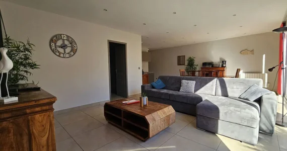 Appart hôtel Pasino n° coin repas à gauche et à l'arrière vue du patio appartement, à droite la partie salon un grand canapé gris un meuble " tiroirs en teck, un écran TV grand écran est posé dessus 2 éléments de décoration un échassier et un poisson en bois