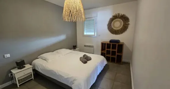 Appart hôtel Pasino n°2 Chambre On y voit un grand lit 160/200 recouvert d'une couette avec housse blanche et ses 2 oreillers et leur taie. 2 draps de douches gris sont posés sur la couette. Deux chevets à tiroir attenants au lit. Côté mur, une fenêtre donnant sur le palier et un meuble de rangement en teck. Décoration exotique.