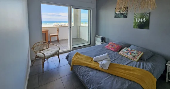 Appart hôtel Pasino n°1 Chambre donnant sur la terrasse vue mer, 2 chevets, 2 lampes de chevet, 2 coussins décoration sur le lit, un plaid jaune, 2 serviettes posées sur le lit. Décorations au mur et plafond. sur la gauche un fauteuil en rotin. A l'arrière de la photo vue sur la terrasse et l'océan