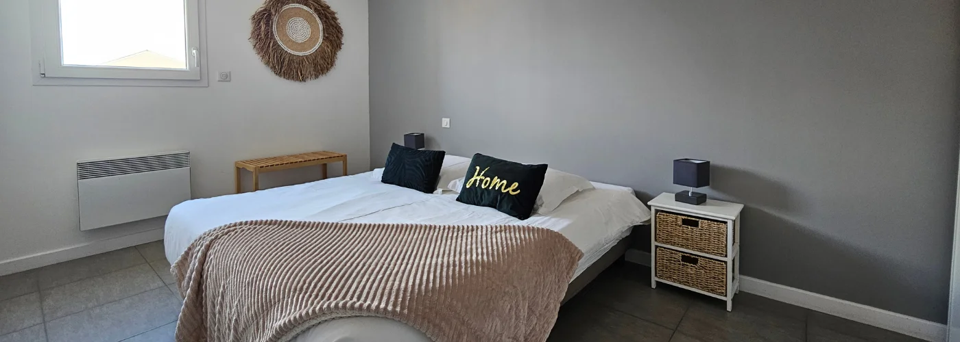 appart hôtel pasino n°6, chambre N°3 murs blancs et gris, une décoration exotique sur le mur blanc dessous un banc en pin, une suspension au plafond, 2 chevets blancs : 2 tiroirs en Osiers plus lampes grises, un lit en 160X200 , 2 oreillers, 2 coussins décoration Vert foncé l'un marqué "Home",