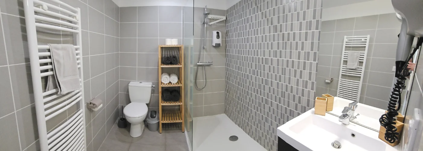 Appart hôtel Pasino n°2 salle de bain avec sa douche italienne, son meuble vasque et son miroir. équipée d'un radiateur sèche-serviette et un sèche cheveux. Une étagère avec 6 draps de douche