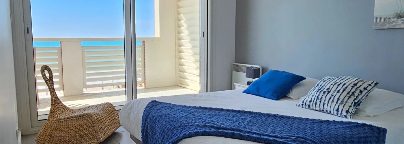 appart hôtel pasino n°5, chambre donnant sur la terrasse vue mer, 2 lampes de chevet, 2 coussins décoration, 1 plaid posé sur le lit. Décorations au mur et plafond. A l'arrière de la photo vue sur la terrasse et l'océan