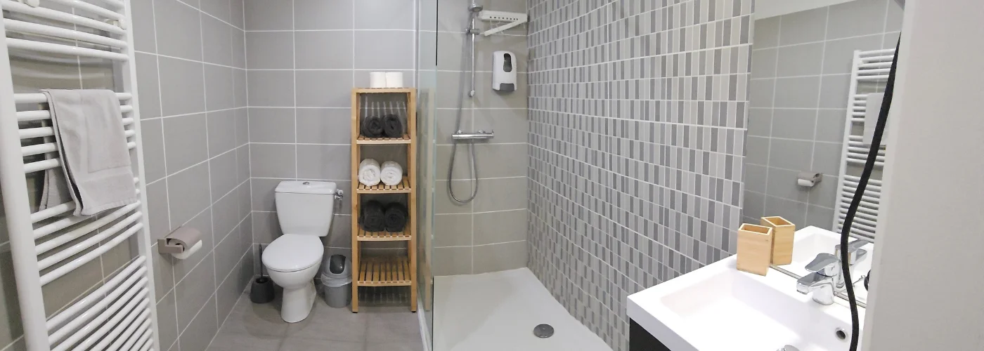 Appart hôtel Pasino n°5 salle de bain avec sa douche italienne, son meuble vasque et son miroir. équipée d'un radiateur sèche-serviette et un sèche cheveux. Une étagère avec 6 draps de douche