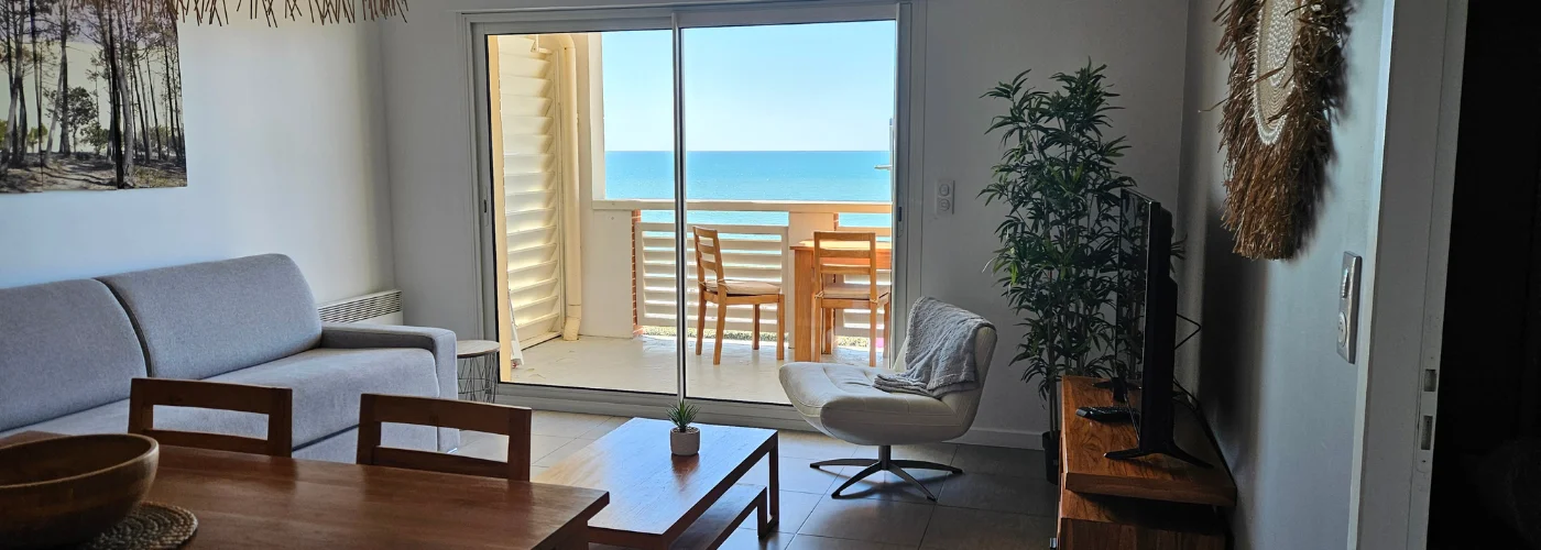 appart hôtel pasino n°5, pièce principale , un canapé gris, des meubles en teck, un écran TV, un fauteuil blanc en cuir Présence d'éléments de décoration. Vue sur la terrasse et l'océan