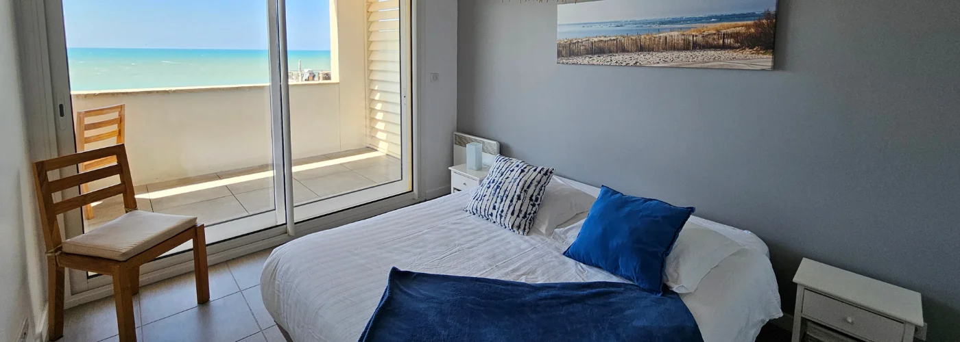 Appart hôtel Pasino N°2, chambre donnant sur la terrasse vue mer, 2 chevets, 2 lampes de chevet, 2 coussins décoration, 1 plaid posé sur le lit. Décorations au mur. A l'arrière de la photo vue sur la terrasse et l'océan