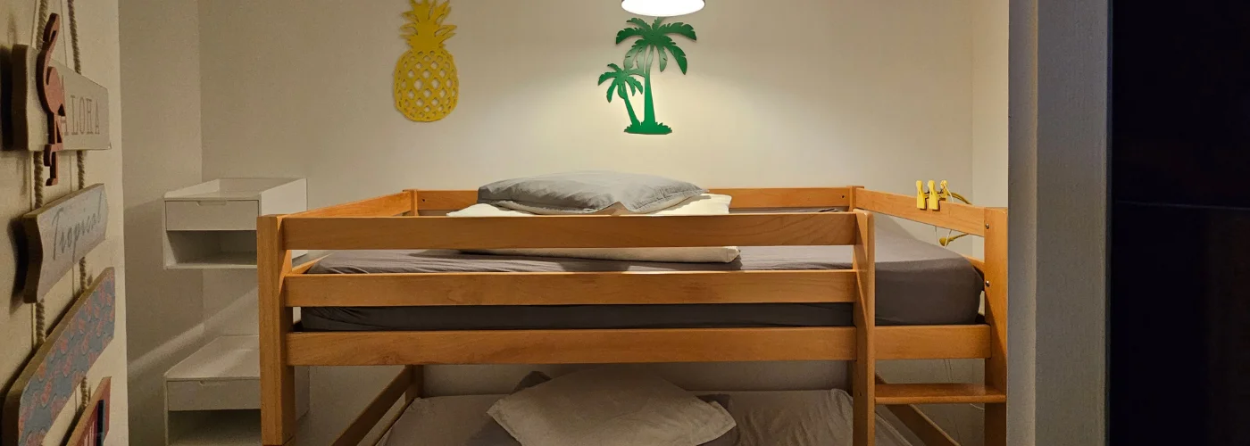 Appart hôtel Pasino N°1, Petite chambre aveugle contient des lits superposés en pin. décorations ananas et palmier au mur Sur chaque lit, Une couette, un oreiller sur un matelas gris. A gauche au mur une table de chevet pour chaque lit