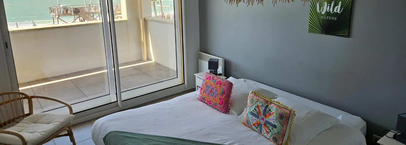 Appart hôtel Pasino n°1 Chambre donnant sur la terrasse vue mer, 2 chevets, 2 lampes de chevet, 2 coussins décoration sur le lit, un plaid vert posées sur le lit. Décorations au mur et plafond. sur la gauche un fauteuil en rotin. A l'arrière de la photo vue sur la terrasse et l'océan