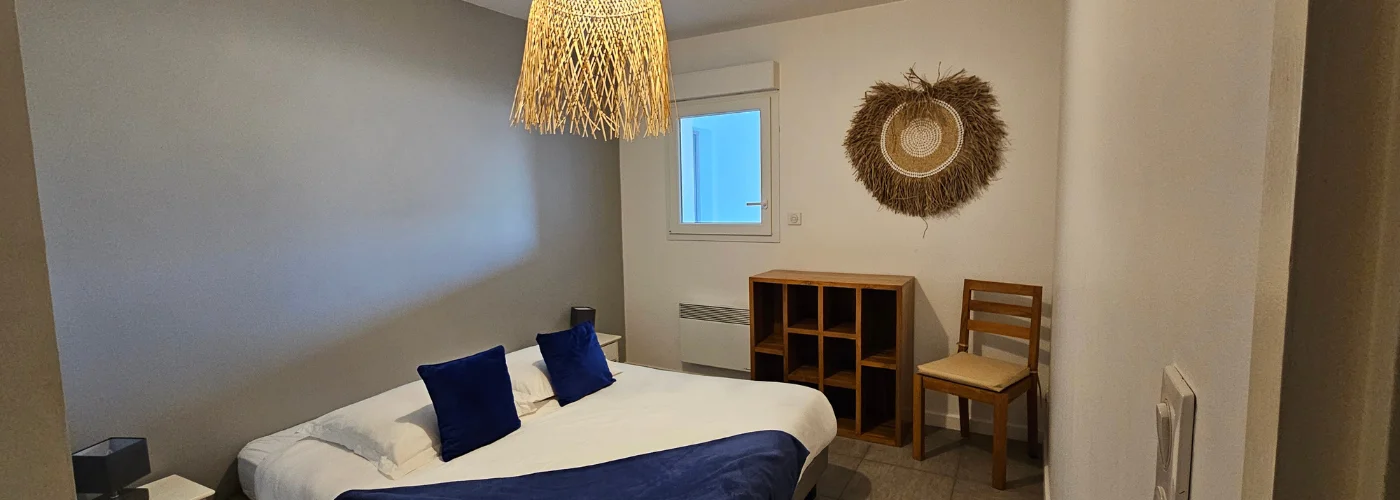 appart hôtel pasino n°2, chambre de l'appartement n°2 de la résidence Pasino. On y voit un grand lit 160/200 recouvert d'une couette avec housse blanche et ses 2 oreillers et leur taie. Un plaid et 2 coussins bleus posés sur la couette. Deux chevets à tiroir attenants au lit. Côté mur, une fenêtre donnant sur le palier et un meuble de rangement en teck. Décoration exotique.
