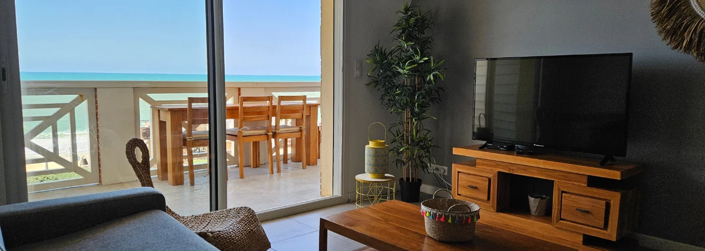 Appart Hôtel Pasino N°4, pièce à vivre de l'appartement numéro 4, elle est lumineuse avec ses murs blancs, A l'arrière la partie salon: sa table de salon en teck , son meuble TV et la TV grand écran. des éléments de décoration au mur, un palmier au fond de la pièce. une baie vitrée donne sur la terrasse vue mer et ses meubles en teck