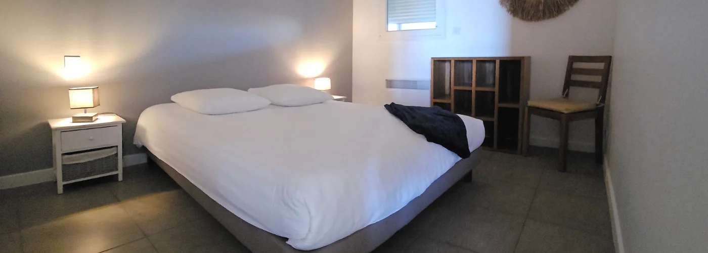 appart hôtel pasino n°5, chambre de l'appartement n°2 de la résidence Pasino. On y voit un grand lit 160/200 recouvert d'une couette avec housse blanche et ses 2 oreillers et leur taie. Un plaid et 2 coussins bleus posés sur la couette. Deux chevets à tiroir attenants au lit. Côté mur, une fenêtre donnant sur le palier et un meuble de rangement en teck. Décoration exotique.
