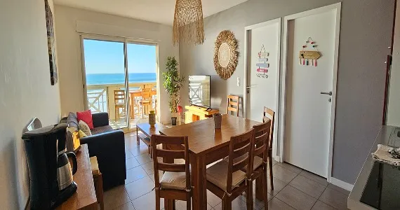 Appart-hotel-appartement-n4-piece-principale-570x300 Appart Hôtel Pasino N°4, la photo est prise à l'intérieur et on découvre le séjour comportant une table en bois et ses 4 chaises, un buffet et un canapé gris. Le sol est carrelé, la décoration est moderne.