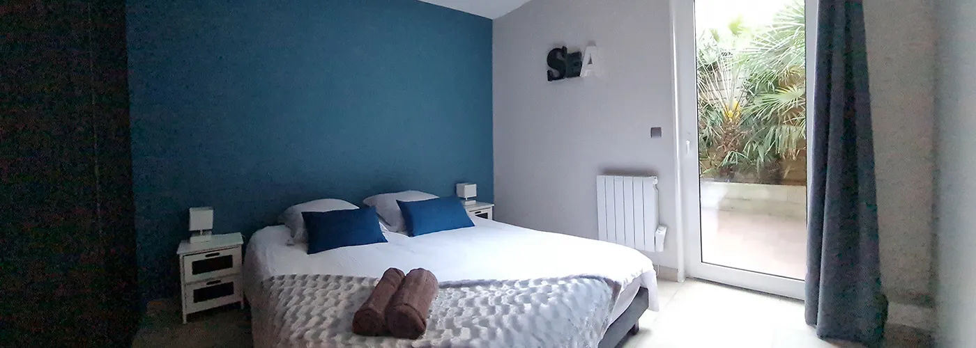 /appart hôtel pasino n°8, chambre aux tons dominante de bleus, au mur une décoration "sea". La chambre est composée d'un grand lit et ses 2 chevets blancs à tiroirs gris et leur lampe. Sur le lit, 2 coussins de décoration bleuet, un plaid gris et ses 2 serviettes grises posées dessus. A l'arrière une porte fenêtre et son rideau occultant bleu un patio et ses palmiers