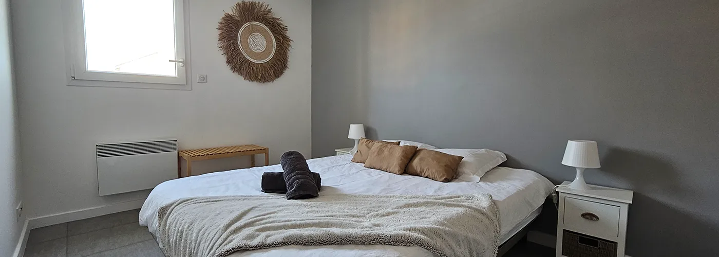/appart hôtel pasino n°3, chambre N°3 murs blancs et gris, une décoration exotique sur le mur blanc dessous un banc en pin, une suspension au plafond, 2 chevet plus lampes, un lit en 160X200 , 2 oreillers, 3 coussins beiges, un plaid et 2 serviettes sur le lit