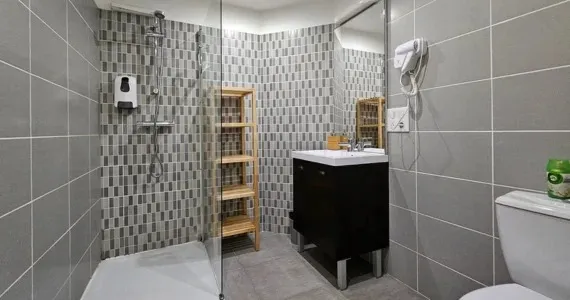 Appart-hotel-location-appartement-n4-salle-de-bain-570x300 Appart hôtel Pasino n°1 Meuble vasque et son miroir, à sa gauche une étagère de rangement en bois et une douche italienne équipée d'une paroi anti projection en verre