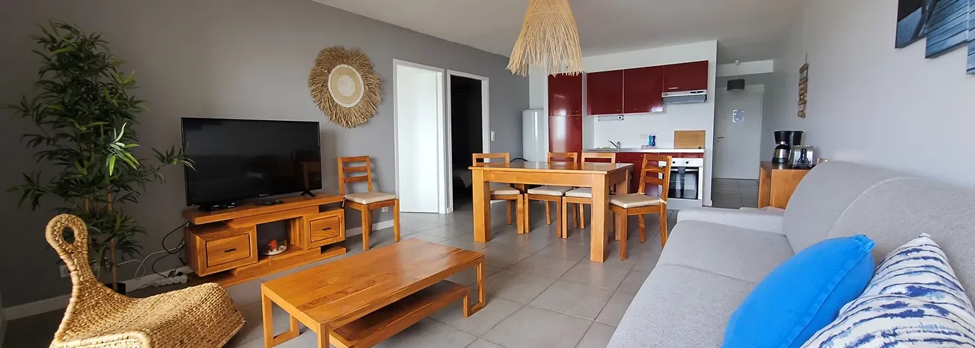 /Appart hôtel Pasino n°2, pièce principale, au premier plan le coin salon et ses meuble en teck, son palmier et sa TV grand écran. et son canapé convertible Au centre une tables et ses chaises et la Kitchenette équipée aux façades rouges. A droite une console .