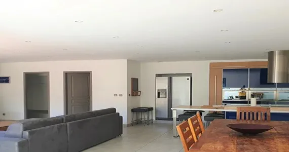 Appart-hotel-residence-pasino-appartement-n7-piece-principale-P-1-redimensionne-taille-570x300 appart hôtel pasino n°7, à louer pour vos vacances. Ici vous pouvez voir la grande pièce à vivre comportant une grande cuisine ouverte toute équipée avec un grand îlot dans les ton bois et bleu, un espace séjour avec une grande table en bois et ses chaises en bois, ainsi qu'un grand canapé d'angle en velours gris anthracite