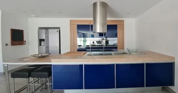 Lacanau-Appart-hotel-residence-pasino-appartement-n7-cuisine-1-570x300 appart hôtel pasino n°7, la grande cuisine aux façades bleues laquées. De nombreux placards de rangement. Un plan de travail couleur bois. sur l'ilot central. Un évier et la table de cuisson sont au centre de celui-ci. au dessus une hotte en inox. A l'arrière, un rectangle de placard couleur bois entoure un ensemble de placard, en haut de fours au centre et à nouveau placard de rangement en bas ainsi que le lave-vaisselle. A gauche le réfrigérateur américain.