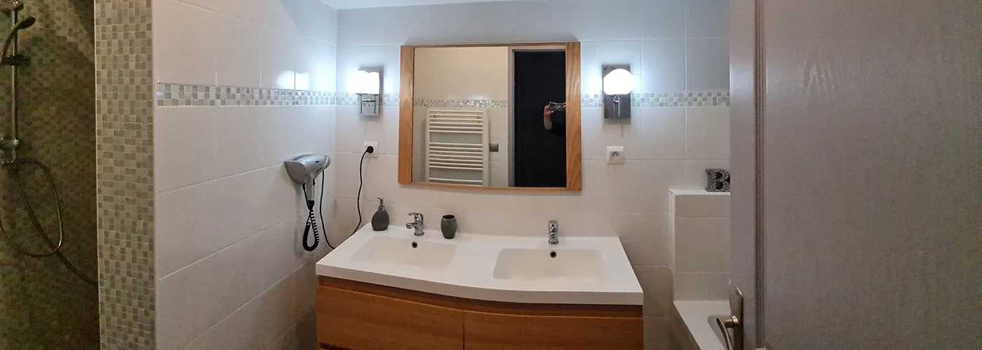 Lacanau-Appart-hotel-residence-pasino-appartement-n7-salle-de-bain appart hôtel pasino n°7, salle de bain, au premier plan, une double vasque, en face, un miroir entouré de bois beige de chaque coté du miroir des appliques argentées avec un globe blanc., juste en dessous une prise électrique. On voit dans le reflet du miroir un radiateur électrique. Sur la paroi du mur, un sèche-cheveux. A gauche la douche italienne et à droite la baignoire. Sous les double vasques, un meuble sous vasque 4 tiroirs en bois clair. Une poubelle est posée au sol.