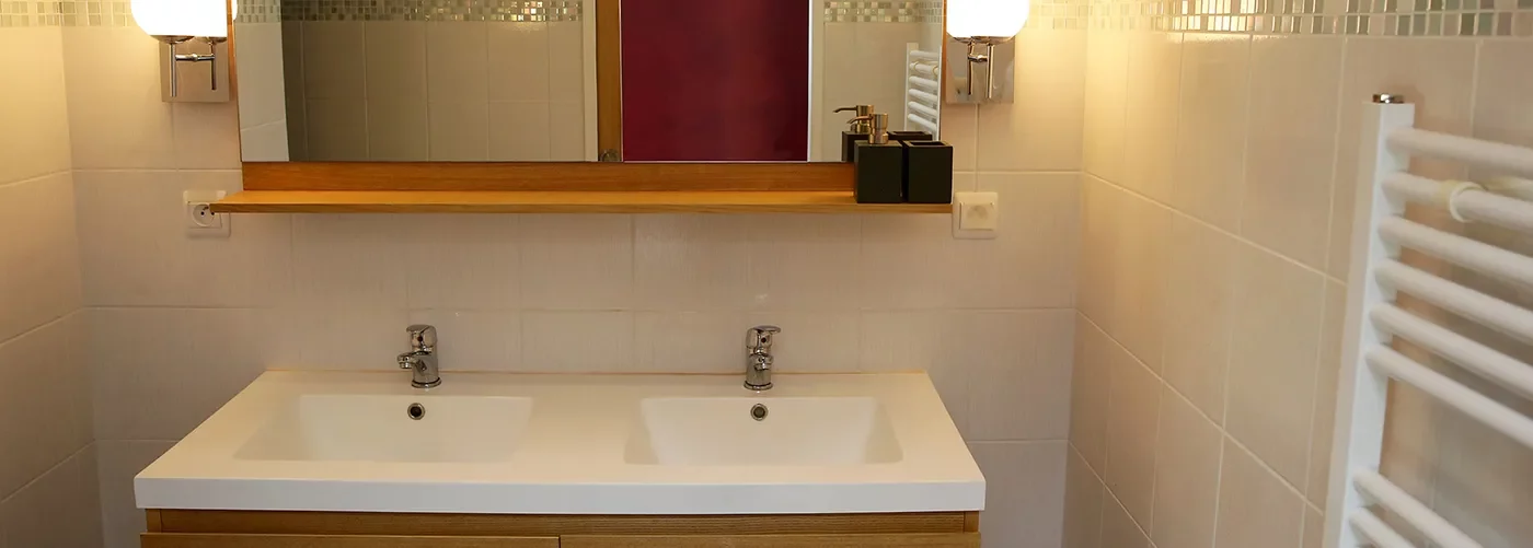Lacanau-Appart-hotel-residence-pasino-appartement-n7-salle-deau appart hôtel pasino n°7, salle de bain, au premier plan, une double vasque, en face, un miroir entouré de bois beige de chaque coté du miroir des appliques argentées avec un globe blanc., juste en dessous une prise électrique. A droite un radiateur électrique. Sous les double vasques, un meuble sous vasque 4 tiroirs en bois clair.