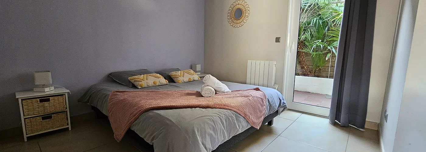 appart hôtel pasino n°8, chambre aux tons dominante de gris, au mur une décoration miroir soleil entouré de rotin. La chambre est composée d'un grand lit et ses 2 chevets blancs à tiroirs en rotin et leur lampe. Sur le lit , 2 coussins de décoration déco ethnique , 2 serviettes blanches posées sur un plaid ROSE. A l'arrière une porte fenêtre et son rideau occultant gris donnant sur un patio et ses palmiers