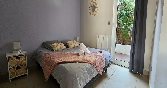 Location-Lacanau-apparthotel-appartement-n8-lacanau-chambre-16taille-570x300 appart hôtel pasino n°8, chambre aux tons dominante de gris, au mur une décoration miroir soleil entouré de rotin. La chambre est composée d'un grand lit et ses 2 chevets blancs à tiroirs en rotin et leur lampe. Sur le lit , 2 coussins de décoration déco ethnique , 2 serviettes blanches posées sur un plaid ROSE. A l'arrière une porte fenêtre et son rideau occultant gris donnant sur un patio et ses palmiers