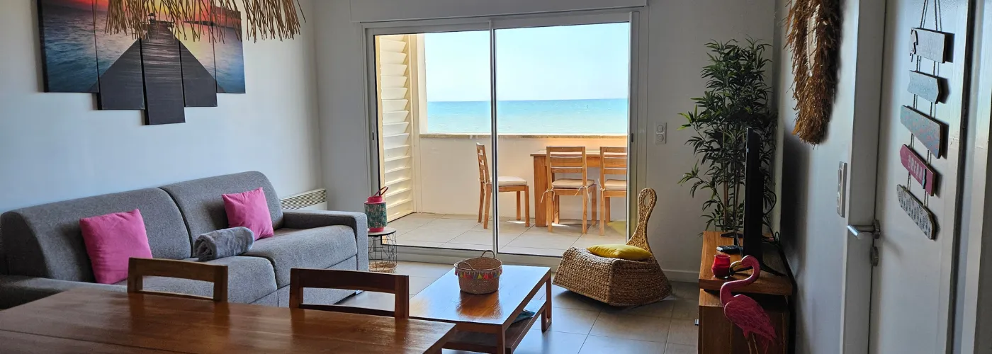 Appart hôtel Pasino n°1 pièce principale , un canapé gris et ses coussins colorés, des meubles en teck, un fauteuil en rotin. Présence d'éléments de décoration aux murs, sur la table de salon et plafond. Au fond de la pièce un palmier. La pièce donne sur une grande terrasse vue mer.