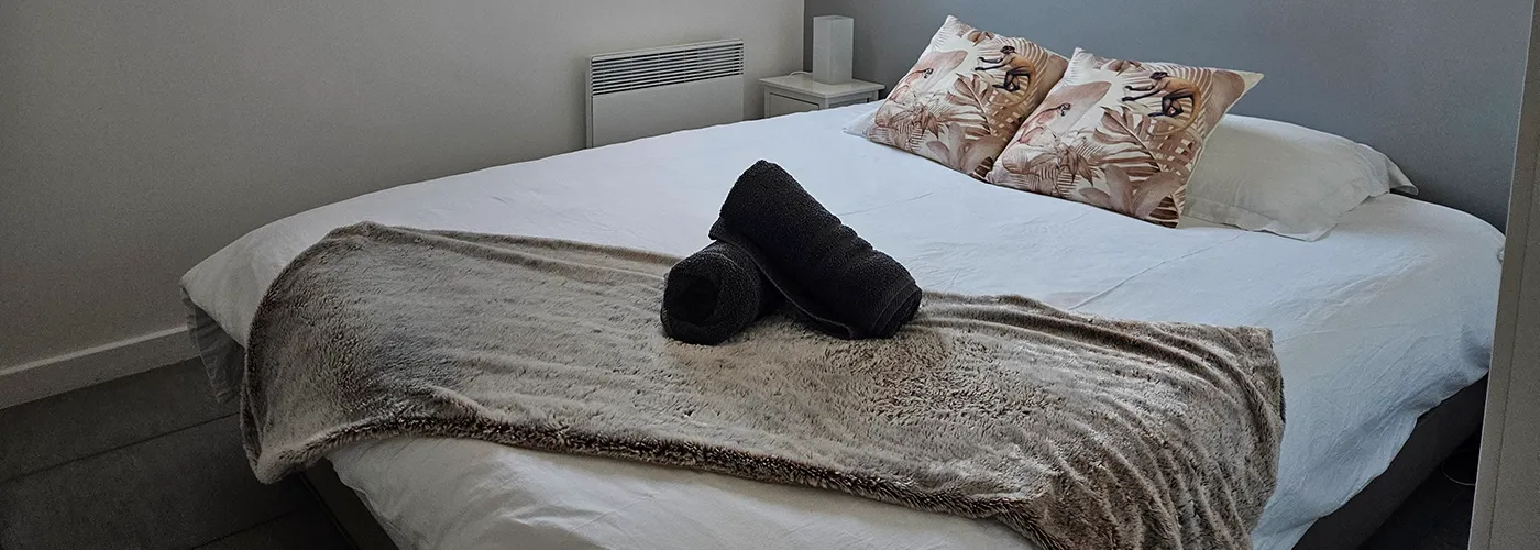 appart hôtel pasino n°3, chambre N°2 une fenêtre sur mur blanc, sur un mur gris, 2 chevets plus lampes, un lit en 160X200 , 2 oreillers, 2 coussins imprimé exotique beige, un plaid et 2 serviettes sur le lit