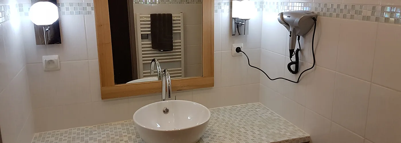 Salle de bain un mélange de petits et moyens carreaux blancs et verts très clair. Au centre un meuble et sa vasque au dessus un miroir, entouré de 2 appliques. Accroché au mur à droite, un sèche cheveu