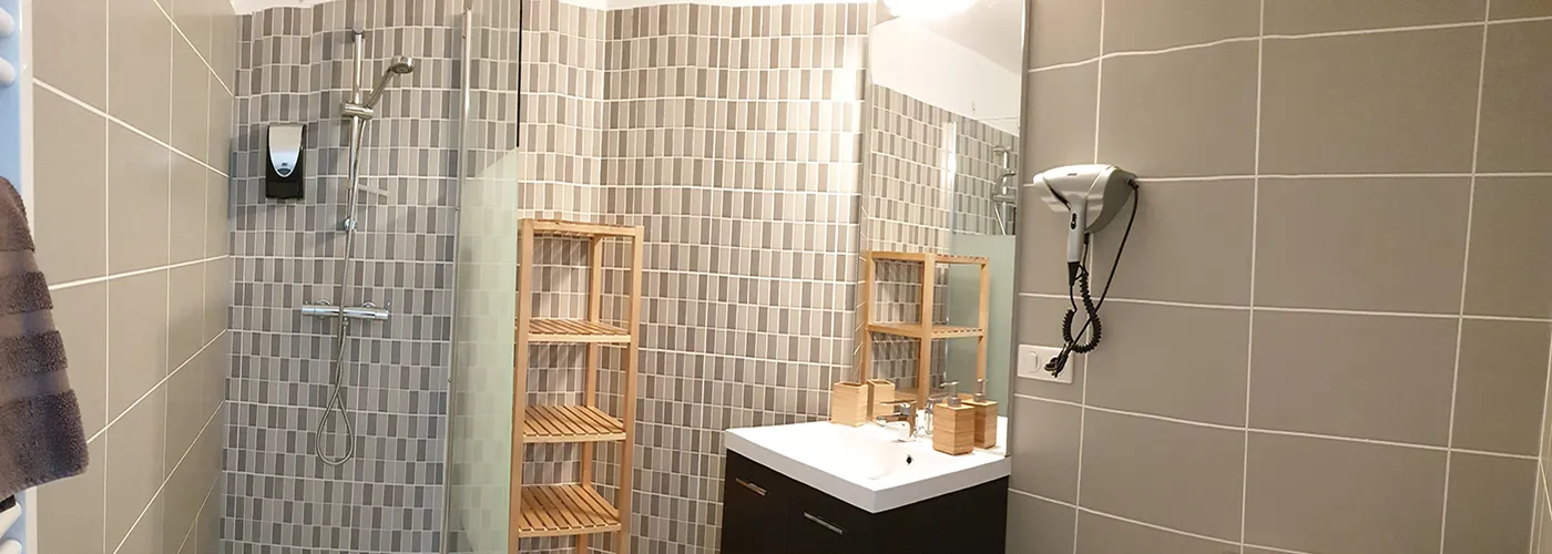 Appart hôtel Pasino n°1 Meuble vasque et son miroir, à sa gauche une étagère de rangement en bois et une douche italienne équipée d'une paroi anti projection en verre