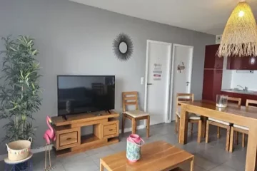 Appart hôtel Pasino n°1 Pièce à vivre, elle est lumineuse avec ses murs blancs,. La pièce est meublée avec des meubles en teck: une table, ses six chaises et leurs coussin d'assise beiges, à gauche une console sur laquelle sont posés un grille pain, une cafetière et une bouilloire. A l'arrière la partie salon : son canapé gris et ses Coussins de décoration jaune et rose, sa table de salon en teck , son meuble TV et la TV grand écran. un fauteuil en rotin. Des éléments de décoration au mur et au plafond, un palmier au fond de la pièce. une baie vitrée donne sur la terrasse vue mer et ses meubles en teck