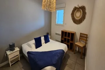 appart hôtel pasino n°2, chambre de l'appartement n°2 de la résidence Pasino. On y voit un grand lit 160/200 recouvert d'une couette avec housse blanche et ses 2 oreillers et leur taie. Un plaid et 2 coussins bleus posés sur la couette. Deux chevets à tiroir attenants au lit. Côté mur, une fenêtre donnant sur le palier et un meuble de rangement en teck. Décoration exotique.