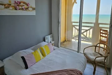 Chambre vue mer, une grand lit recouvert d'une couette et ses oreillers blancs.des coussins colorés et un plaide complete le tout. Un fauteuil en osier et son coussin est pres de la baie vitree un tableau est posé sur le mur gris au dessus du lit. La vue mer donnes sur un océan aux couleurs estivales