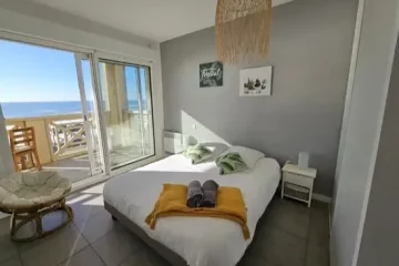 Appart Hôtel Pasino N°4, chambre appartement donnant sur la terrasse vue mer, 2 chevets, 2 lampes de chevet, 2 coussins décoration sur le lit, un plaid jaune, 2 serviettes posées sur le lit. Décorations au mur et plafond. sur la gauche un fauteuil en rotin. A l'arrière de la photo vue sur la terrasse et l'océan