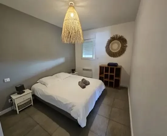 Appart hôtel Pasino n°2 Chambre. On y voit un grand lit 160/200 recouvert d'une couette avec housse blanche et ses 2 oreillers et leur taie. 2 draps de douches gris sont posés sur la couette. Deux chevets à tiroir attenants au lit. Côté mur, une fenêtre donnant sur le palier et un meuble de rangement en teck posé dessus Un plaid bleu. Décoration exotique.