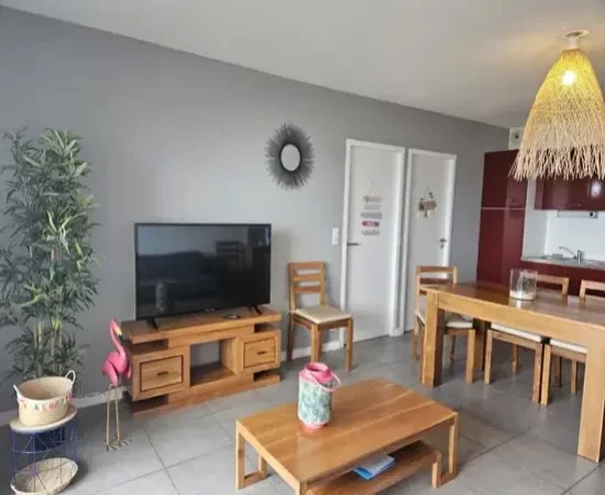 Appart hôtel Pasino n°1 Pièce à vivre, elle est lumineuse avec ses murs blancs,. La pièce est meublée avec des meubles en teck: une table, ses six chaises et leurs coussin d'assise beiges, à gauche une console sur laquelle sont posés un grille pain, une cafetière et une bouilloire. A l'arrière la partie salon : son canapé gris et ses Coussins de décoration jaune et rose, sa table de salon en teck , son meuble TV et la TV grand écran. un fauteuil en rotin. Des éléments de décoration au mur et au plafond, un palmier au fond de la pièce. une baie vitrée donne sur la terrasse vue mer et ses meubles en teck