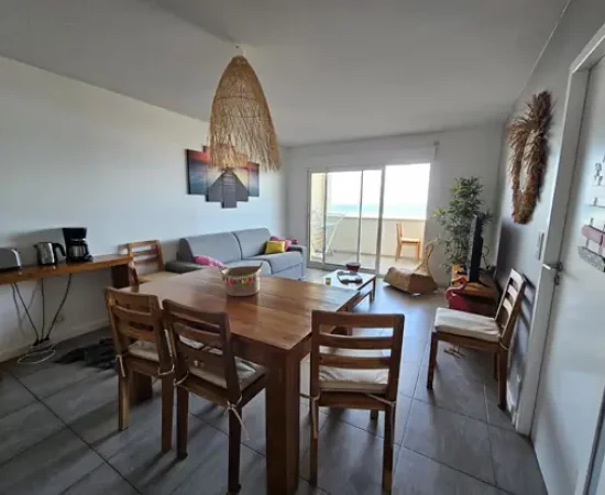 Appart hôtel Pasino n°1 Pièce à vivre, elle est lumineuse avec ses murs blancs,. La pièce est meublée avec des meubles en teck: une table, ses six chaises et leurs coussin d'assise beiges, à gauche une console sur laquelle sont posés un grille pain, une cafetière et une bouilloire. A l'arrière la partie salon : son canapé gris et ses Coussins de décoration colorés, sa table de salon en teck , son meuble TV et la TV grand écran, un fauteuil en rotin. Des éléments de décoration au mur, sur les 2 portes et au plafond, un palmier au fond de la pièce. une baie vitrée donne sur la terrasse vue mer et ses meubles en teck