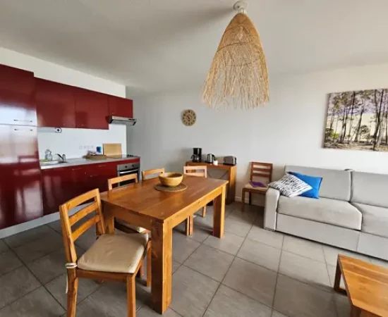 appart hôtel pasino n°5, pièce à vivre de l'appartement de la résidence Pasino. Au 1er plan on peut voir une table en teck et ses quatre chaises. On peut voir la canapé convertible au fond vers la droite, à sa gauche une console en teck. Sur la gauche, la cuisine laquée rouge comporte une plaque vitrocéramiques 3 feux, un four en dessous, un réfrigérateur, de grands placards et 1 évier