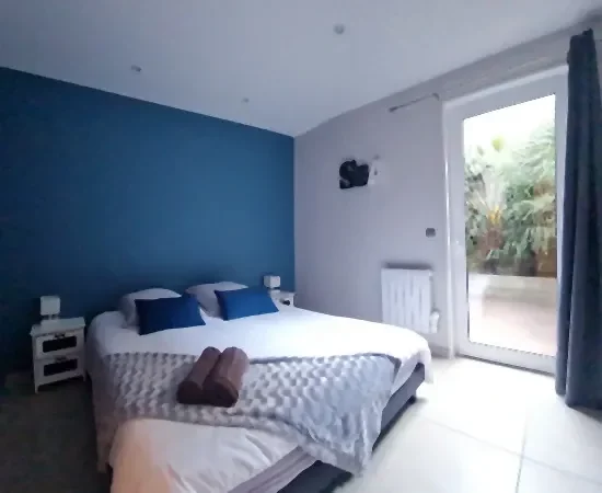 appart hôtel pasino n°8, chambre aux tons dominante de bleus, au mur une décoration "sea". La chambre est composée d'un grand lit et ses 2 chevets blancs à tiroirs gris et leur lampe. Sur le lit 2 coussins de décoration bleuet, un plaid gris et ses 2 serviettes grises posées dessus. A l'arrière une porte fenêtre et son rideau occultant bleu un patio et ses palmiers