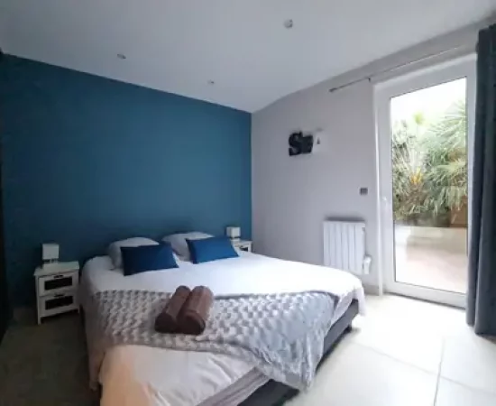 appart hôtel pasino n°8, chambre. Les tons sont en dominante de bleus, au mur une décoration "sea". La chambre est composée d'un grand lit et ses 2 chevets blancs à tiroirs gris et leur lampe. Sur le lit 2 coussins de décoration bleuet, un plaid gris et ses 2 serviettes grises posées dessus. A l'arrière une porte fenêtre et son rideau occultant bleu un patio et ses palmiers
