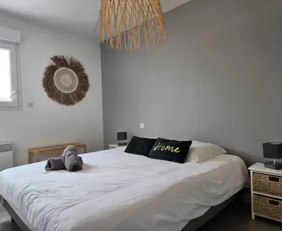 appart hôtel pasino n°6, chambre N°3 murs blancs et gris, une décoration exotique sur le mur blanc dessous un banc en pin, une suspension au plafond, 2 chevets blancs : 2 tiroirs en Osiers plus lampes grises, un lit en 160X200 , 2 oreillers, 2 coussins décoration Vert foncé l'un marqué "Home", 2 serviettes sur le lit