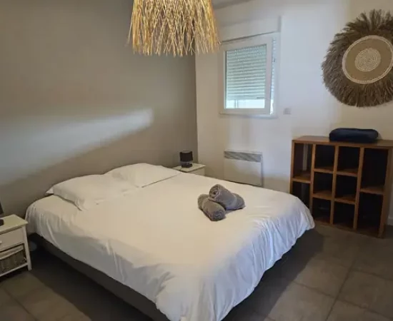 Appart hôtel Pasino N°2, seconde chambre de l'appartement n°2 de la résidence Pasino. On y voit un grand lit 160/200 recouvert d'une couette avec housse blanche et ses 2 oreillers et leur taie. 2 draps de douches gris sont posés sur la couette. Deux chevets à tiroir attenants au lit. Côté mur, une fenêtre donnant sur le palier et un meuble de rangement en teck. Décoration exotique.