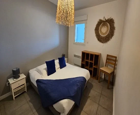 appart hôtel pasino n°2, chambre de l'appartement n°2 de la résidence Pasino. On y voit un grand lit 160/200 recouvert d'une couette avec housse blanche et ses 2 oreillers et leur taie. Un plaid et 2 coussins bleus posés sur la couette. Deux chevets à tiroir attenants au lit. Côté mur, une fenêtre donnant sur le palier et un meuble de rangement en teck. Décoration exotique.