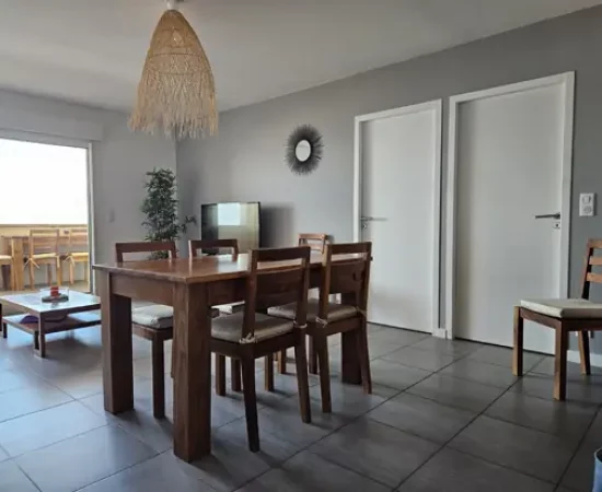 Appart hôtel Pasino N°2, pièce à vivre de l'appartement numéro 2, elle est lumineuse avec ses murs blancs, elle est meublée avec des meubles en teck: une table, ses six chaises et leurs coussin d'assise beiges. A l'arrière la partie salon: sa table de salon en teck , son meuble TV et la TV grand écran. des éléments de décoration au mur, un palmier au fond de la pièce. une baie vitrée donne sur la terrasse vue mer et ses meubles en teck