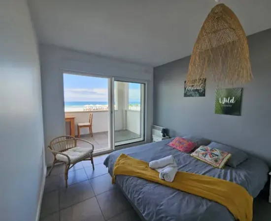 Appart hôtel Pasino n°1 Chambre donnant sur la terrasse vue mer, 2 chevets, 2 lampes de chevet, 2 coussins décoration sur le lit, un plaid jaune, 2 serviettes posées sur le lit. Décorations au mur et plafond. sur la gauche un fauteuil en rotin. A l'arrière de la photo vue sur la terrasse et l'océan